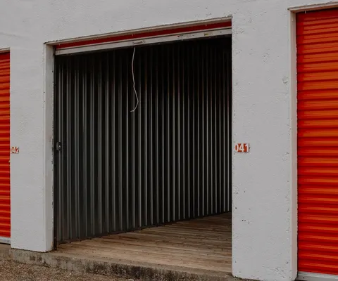 An open drive-up storage unit with a wooden floor between two closed orange roll-up doors