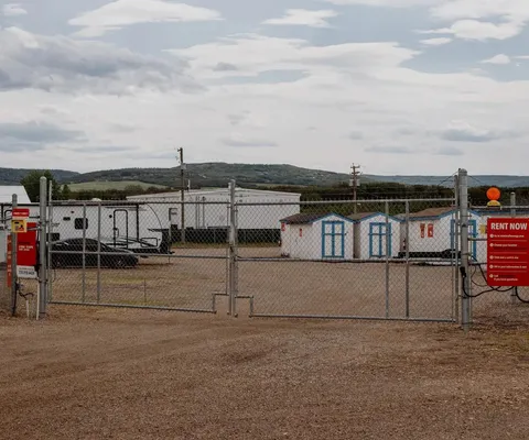 A gated chain-link fence opens to a gravel storage lot with sheds, trailers, and outdoor signage