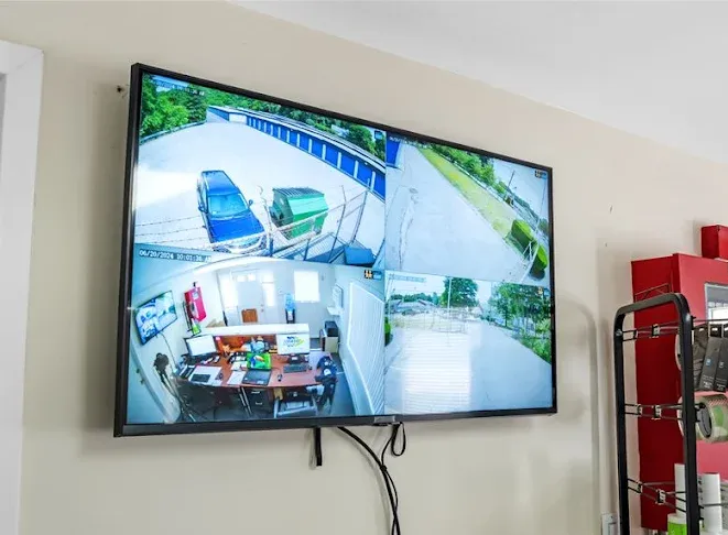 A wall-mounted security monitor displaying four split-screen video feeds of the storage facility office and grounds