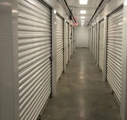 Well-lit indoor storage facility hallway with white corrugated roll-up doors and concrete flooring