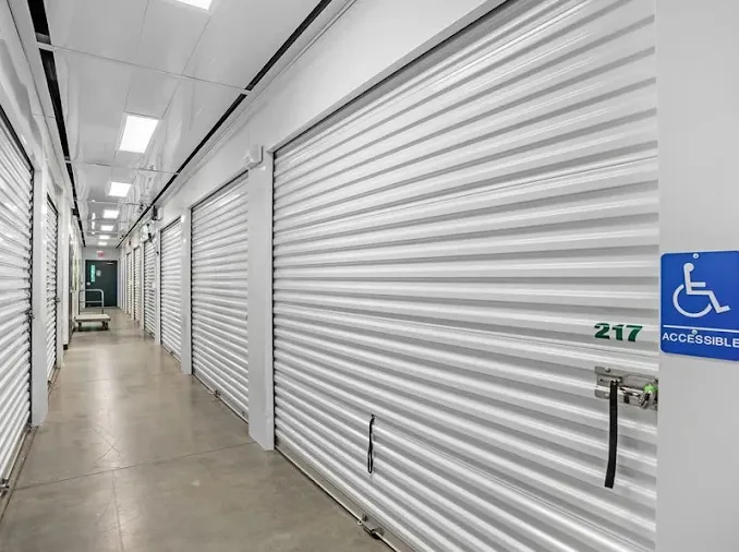 Indoor storage hallway with white roll-up doors, concrete floors, overhead lighting, and an accessible unit sign