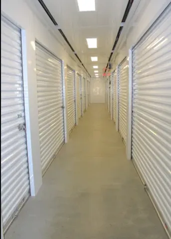 A brightly lit indoor hallway lined with white corrugated metal self-storage unit doors on both sides