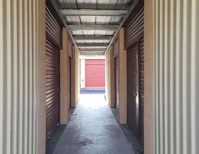 Hallway of covered outdoor storage units in Cohoes at 50 Oliver St, with brownish-red roll-up doors and a corrugated metal ceiling