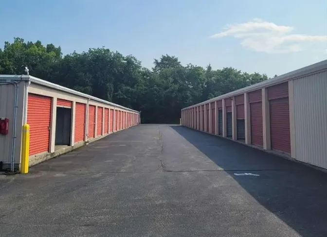 Outdoor drive-up self-storage units with red roll-up doors and a wide asphalt driveway between long buildings