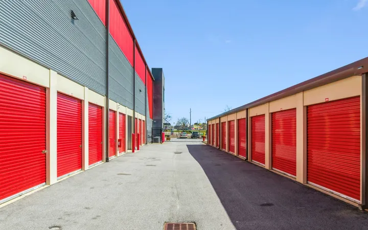 Wide asphalt driveway between drive-up storage units in North Augusta on Cork Tree Bnd with red roll-up doors leading toward a gated entrance under clear skies