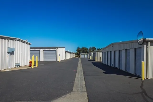 Outdoor drive-up self-storage units in Woodland on Matmor Rd with gray roll-up doors and yellow bollards under a blue sky