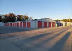 Drive-up outdoor self-storage buildings with red roll-up doors and a wide paved driveway under clear sky