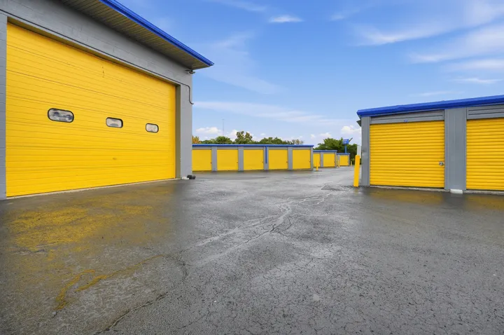 Drive-up storage units in Albany, NY, with yellow roll-up doors and blue trim next to a large warehouse storage unit along an asphalt driveway under a blue sky