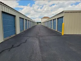 Paved drive aisle between outdoor storage buildings with tan siding and blue roll-up doors under a cloudy sky