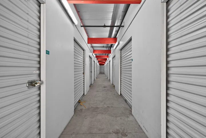 Well-lit hallway of indoor climate-controlled storage in Fairfield on Lopes Rd, featuring white roll-up doors, concrete flooring, and red ceiling beams