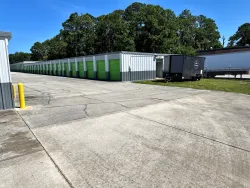 Long row of outdoor drive-up storage units with green doors bordering a paved lot beside trees