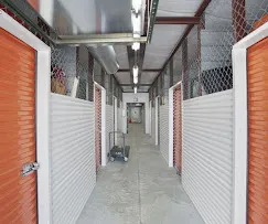 Hallway of interior climate-controlled storage in Little River, SC, with orange roll-up doors, white siding, mesh ceilings, and a moving cart