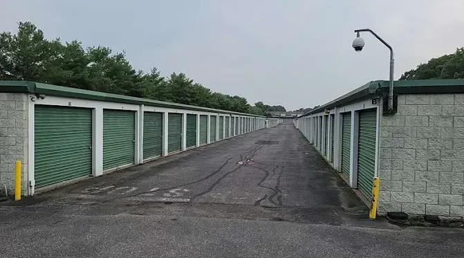 Outdoor drive-up storage units in Holtsville, NY, with green roll-up doors and a security camera overlooking a wide asphalt driveway