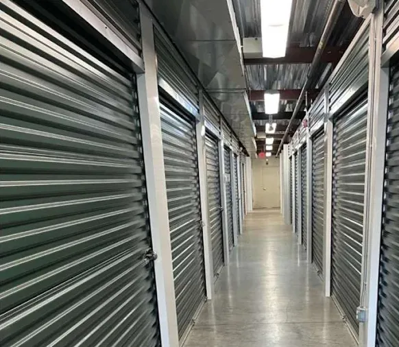 Well-lit indoor hallway lined with green metal roll-up self-storage unit doors on both sides