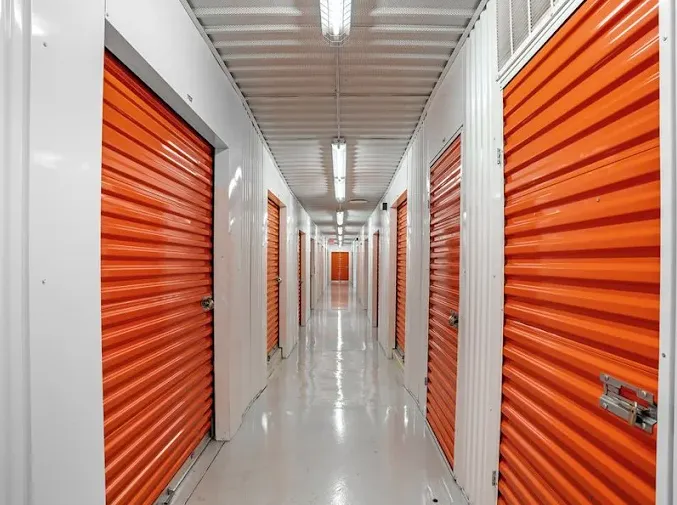 Brightly lit indoor climate-controlled self-storage hallway featuring rows of orange roll-up unit doors and white walls