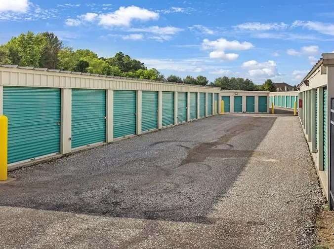 Rows of drive-up self-storage units with green roll-up doors along a paved outdoor driveway under a blue sky