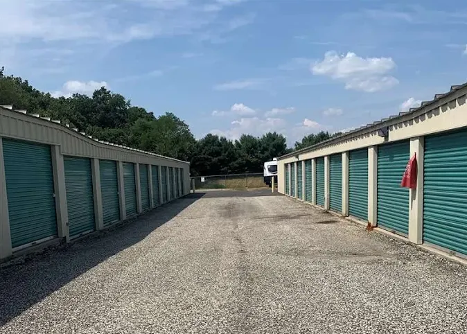 Outdoor drive-up storage units with green roll-up doors line a paved drive aisle under a blue sky