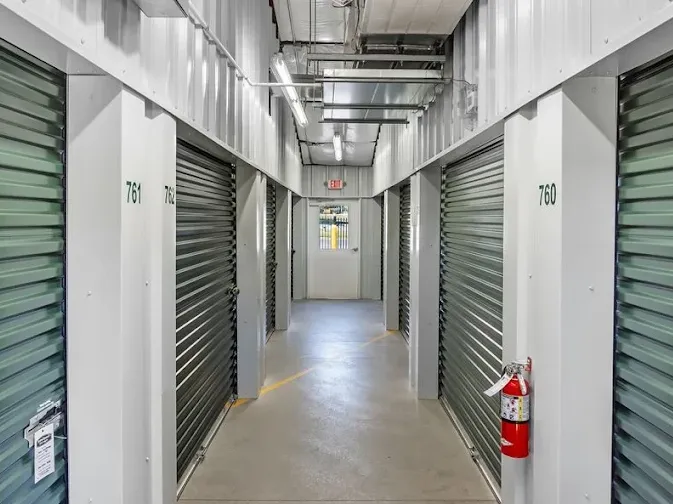 Indoor storage hallway with numbered rolling doors, fluorescent lighting, a fire extinguisher, and an exit door