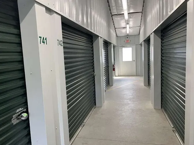 Indoor self-storage hallway with green corrugated roll-up doors, numbered units, and an exit door at the end