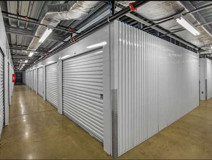 Climate-controlled storage hallway with white roll-up metal doors and exposed ceiling pipes and vents