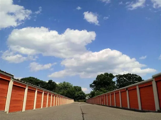 Paved driveway between two long rows of outdoor storage units in Westville, NJ, with orange roll-up doors under a blue sky