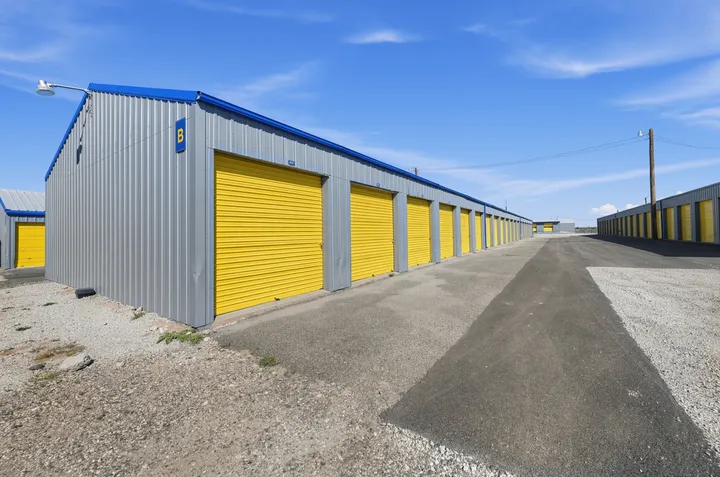 Paved driveway between two rows of gray outdoor drive-up storage units in Laramie, with yellow roll-up doors under a blue sky