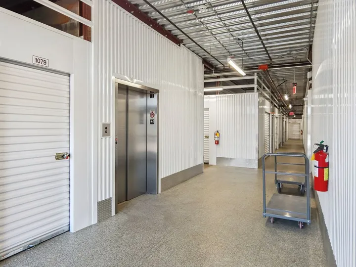 Brightly lit hallway of climate-controlled storage with white corrugated walls, an elevator, and gray moving carts