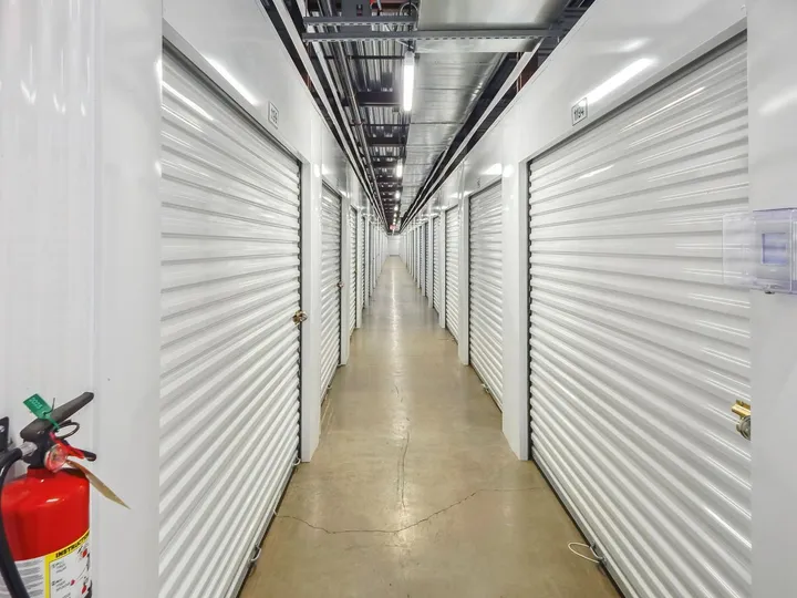 Well-lit interior hallway of a self-storage facility with white roll-up doors and a fire extinguisher