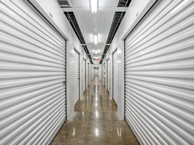 Brightly lit indoor hallway featuring rows of white corrugated roll-up storage unit doors and polished floors