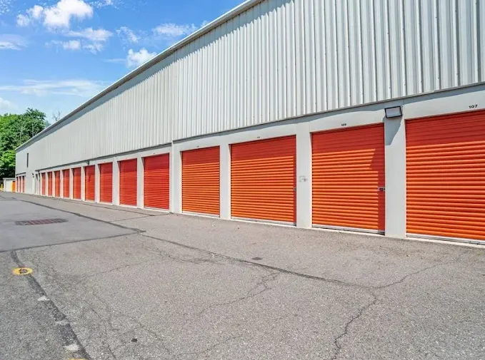 A long row of outdoor drive-up storage units with orange roll-up doors and asphalt access path