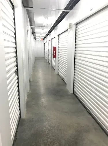 Indoor self-storage hallway with white roll-up doors, concrete flooring, and overhead lighting along a narrow corridor