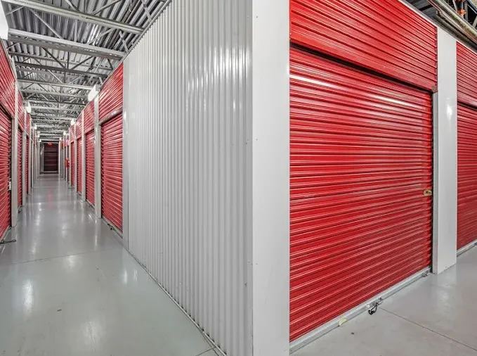 Long indoor climate-controlled storage unit hallway featuring red roll-up storage unit doors and white corrugated metal walls