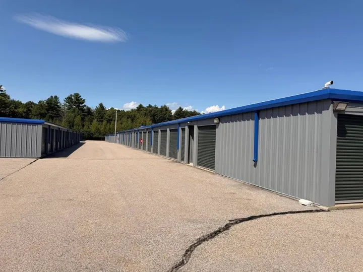 A wide paved driving aisle between rows of gray drive-up storage units in Coventry, RI, with green roll-up doors
