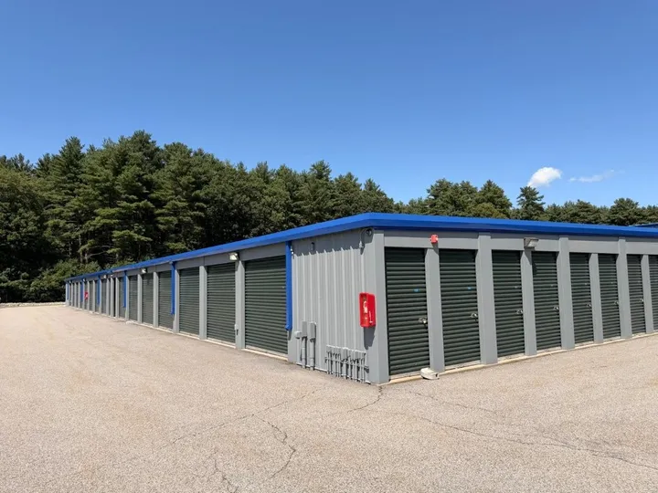 Exterior row of gray drive-up storage units in Coventry on 1185 Tiogue Ave, with green roll-up doors and blue trim under a blue sky