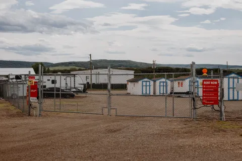 A gated chain-link fence opens to a gravel storage lot with sheds, trailers, and outdoor signage