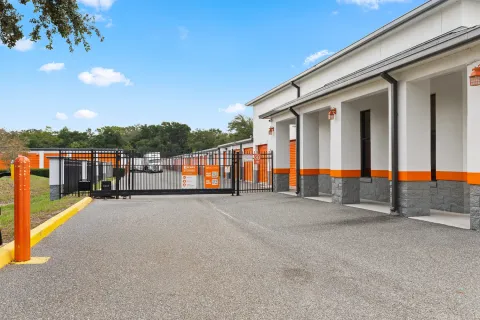 A black metal security gate leads to outdoor storage units with orange roll-up doors and white exteriors
