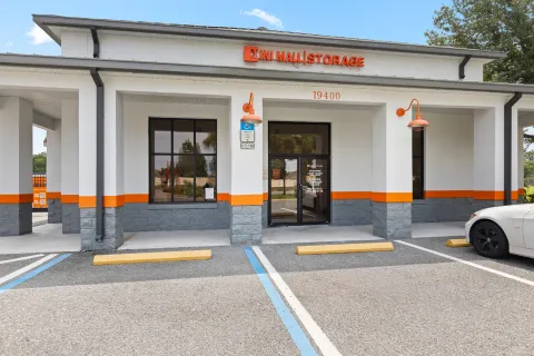A white Mini Mall Storage office building with glass doors, orange accents, and a paved parking lot