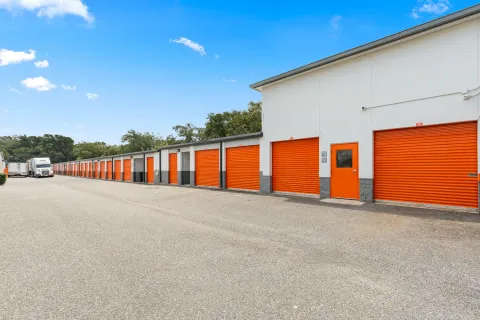 A row of white outdoor self-storage units with orange roll-up doors and a paved driving aisle