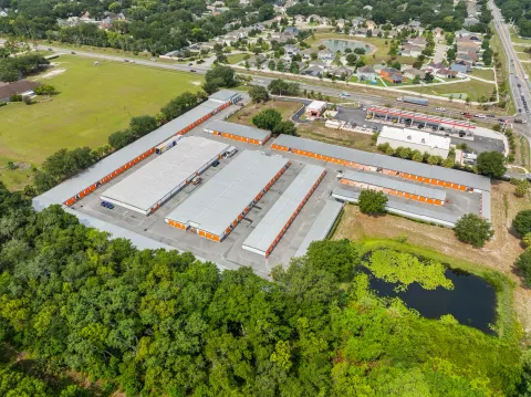 High-angle aerial view of a self-storage facility with orange roll-up doors surrounded by trees and water
