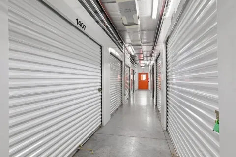 A bright indoor storage hallway with white corrugated metal roll-up doors and a distant orange exit door