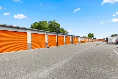 Outdoor drive-up self-storage units with orange roll-up doors and a paved asphalt driveway under a blue sky