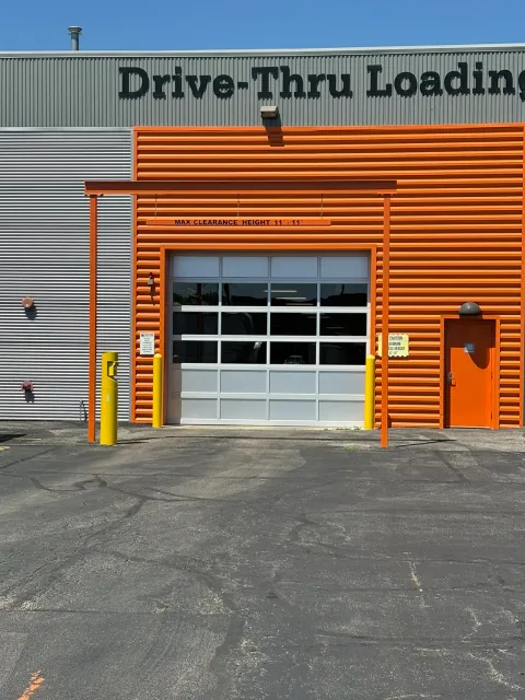 A glass bay door at a drive-thru loading area with orange corrugated metal siding and yellow bollards