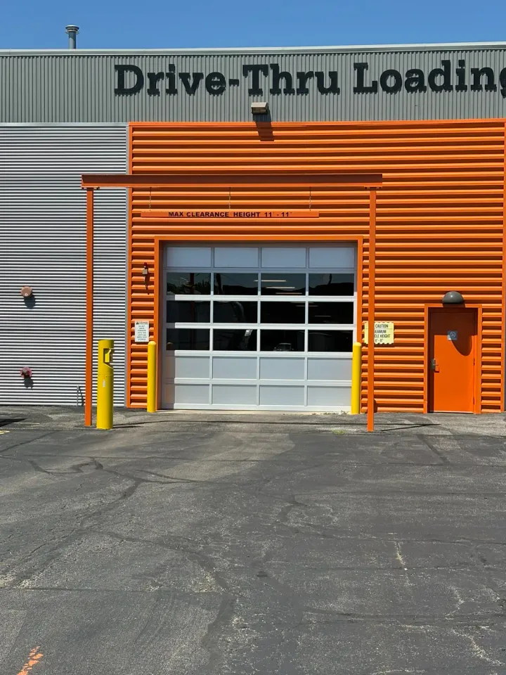 A glass bay door at a drive-thru loading area with orange corrugated metal siding and yellow bollards