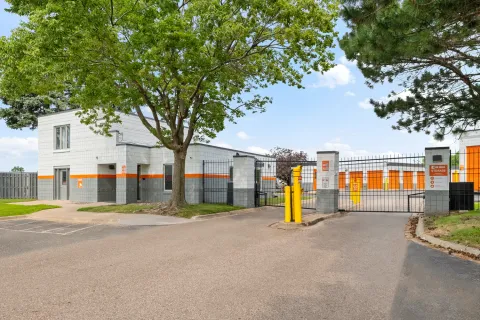 A gated entrance to a storage facility with a stone office building and orange roll-up doors