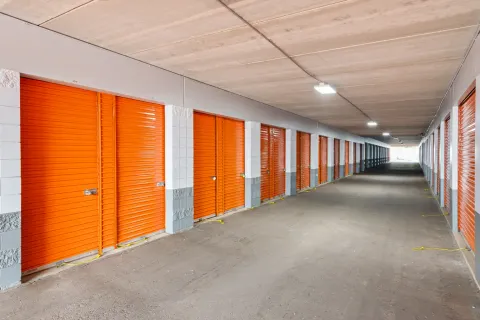 A long interior hallway featuring bright orange roll-up storage unit doors and gray concrete floors