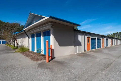 Exterior view of storage unit building with blue roll-up doors and orange entry doors under a clear sky
