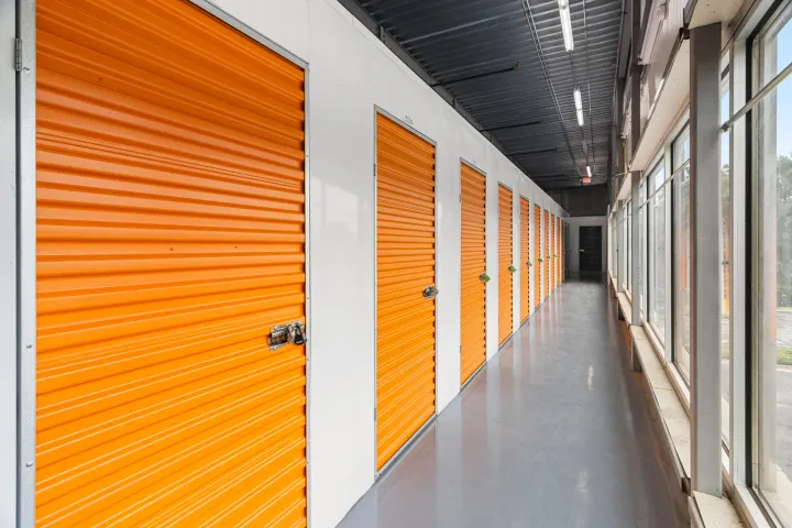 Indoor storage hallway featuring a row of orange roll-up doors and large windows on the right