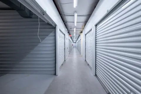 Brightly lit interior hallway with white corrugated metal roll-up storage unit doors and overhead fluorescent lighting