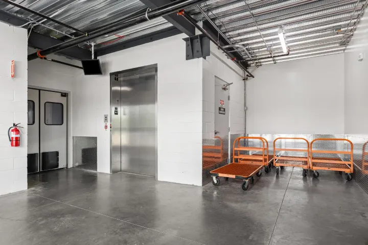 Indoor storage loading area featuring a silver elevator door and several orange flatbed moving carts