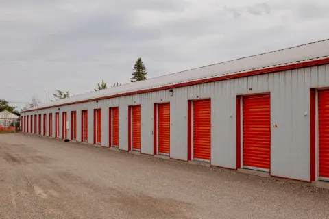 Row of outdoor drive-up storage units with orange roll-up doors and a gravel driveway